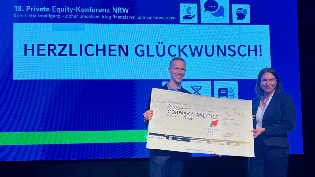 Zwei Personen stehen auf einer Bühne und halten einen großen Scheck über 3000 Euro in den Händen. Im Hintergrund ist ein Bildschirm mit der Aufschrift “18. Private Equity-Konferenz NRW” und “HERZLICHEN GLÜCKWUNSCH” zu sehen.