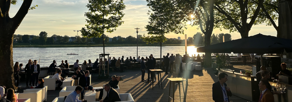 Eine Gruppe von Menschen sitzt und steht auf einer Terrasse am Flussufer bei Sonnenuntergang. Die Szene ist von Bäumen gesäumt, und im Hintergrund ist der Fluss mit einigen Booten und dem gegenüberliegenden Ufer zu sehen. Die Menschen unterhalten sich in kleinen Gruppen, während die Sonne tief am Himmel steht und ein warmes Licht auf die Szene wirft.