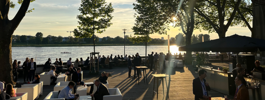 Eine Gruppe von Menschen sitzt und steht auf einer Terrasse am Flussufer bei Sonnenuntergang. Die Szene ist von Bäumen gesäumt, und im Hintergrund ist der Fluss mit einigen Booten und dem gegenüberliegenden Ufer zu sehen. Die Menschen unterhalten sich in kleinen Gruppen, während die Sonne tief am Himmel steht und ein warmes Licht auf die Szene wirft.